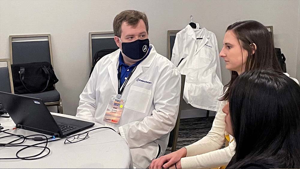 Blog - Man with Mask and Lab Coat on Speaking with Other Professionals While Sitting at his Desk with His Laptop Open