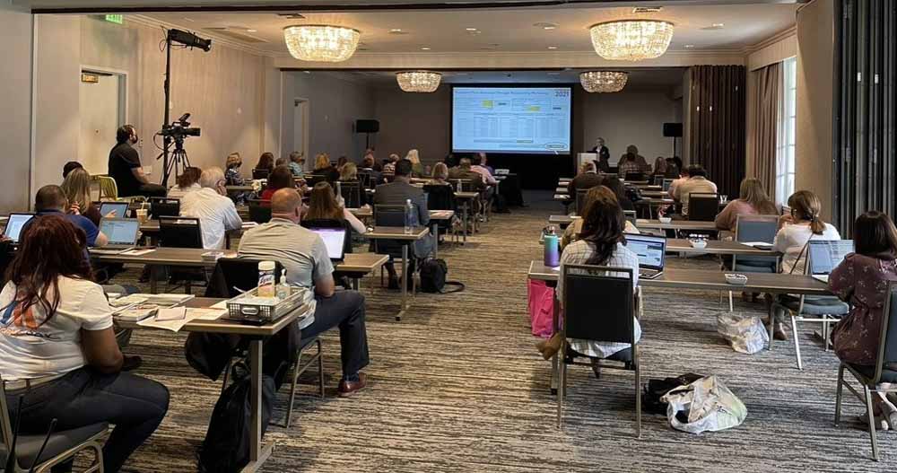Blog - Professionals Sitting with Laptops Open in Front of a Projector and Listening at the 2021 HUG National Conference